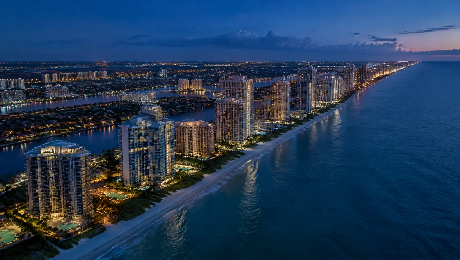 Aerial view at dusk of lit condominium towers along a Florida Atlantic coastline