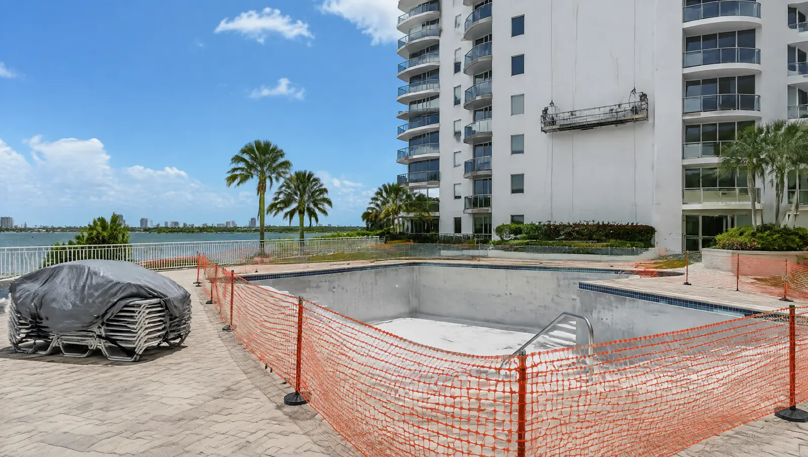 Drained condominium pool fenced off with orange safety netting, covered lounge chairs and an empty work platform on the tower